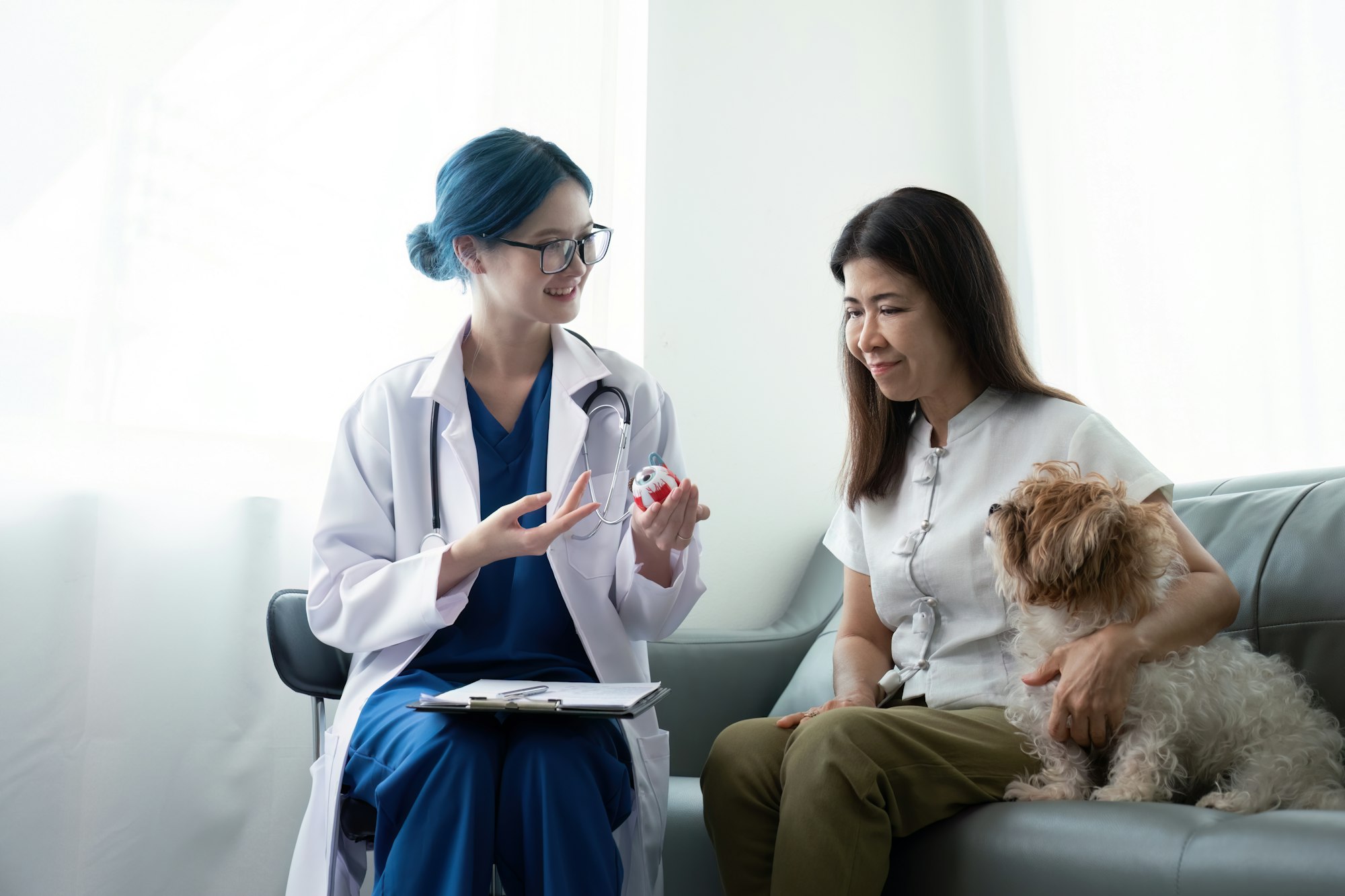 Elderly woman with dog sitting by desk in front of asian woman veterinary at vet clinic